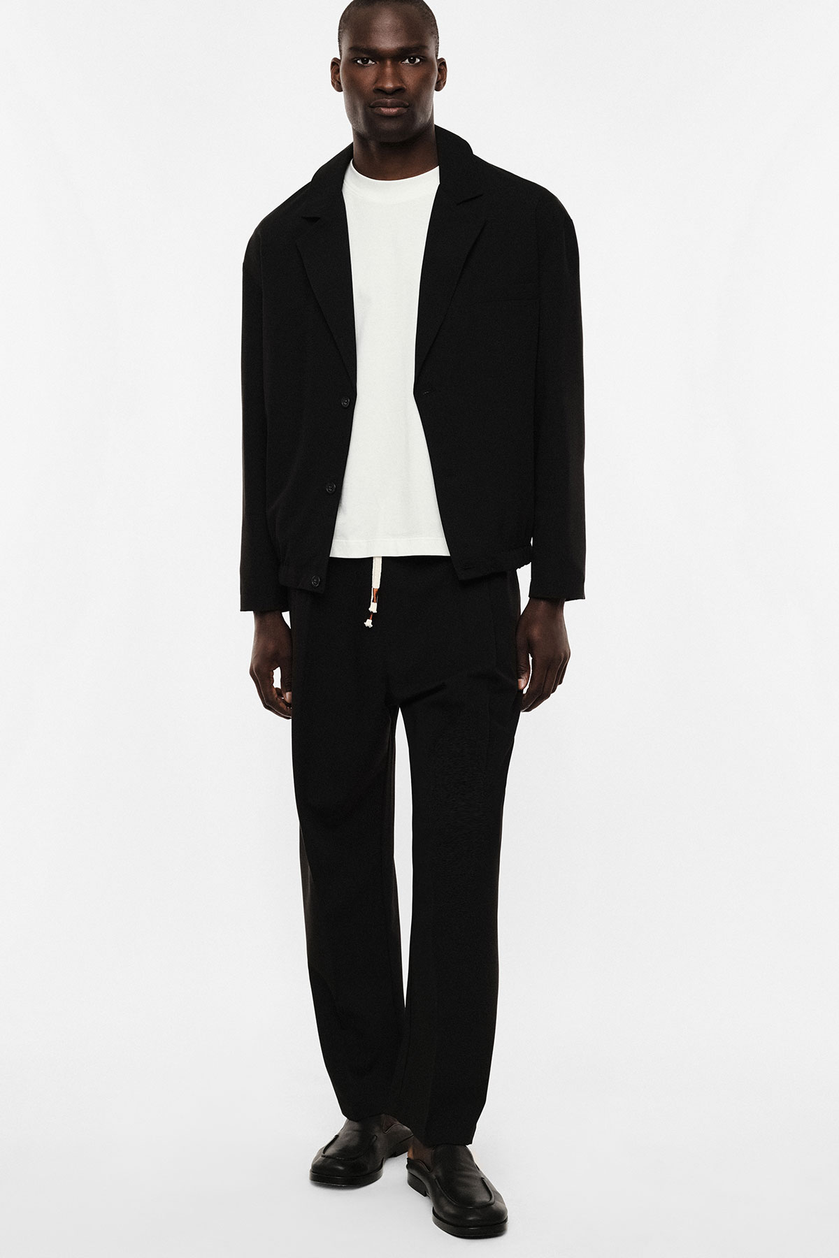 Full-length studio photo of a male model wearing a black jacket and trousers, white T-shirt and black loafers