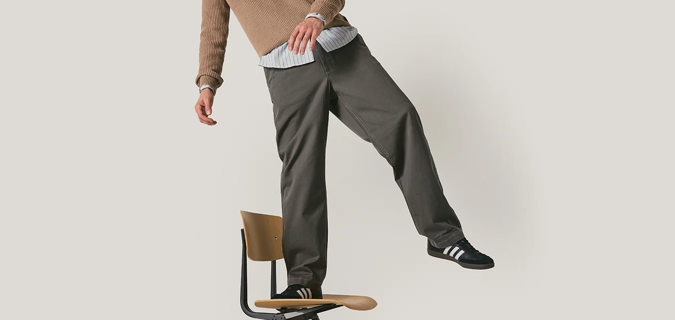 Pantaloni grigi con una vestibilità rilassata, cuciture dettagliate, indossati con un maglione beige a trama e scarpe nere con strisce bianche. In piedi su una sedia.
