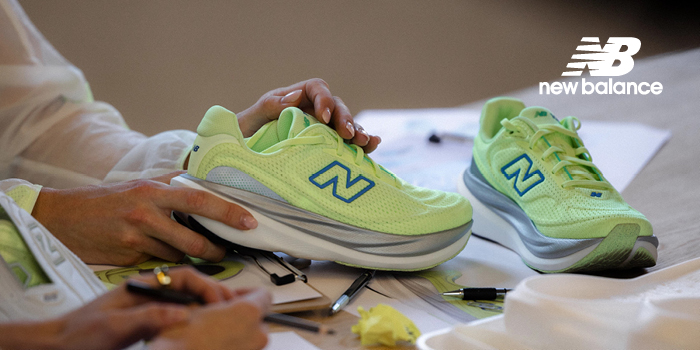 Hands holding neon green New Balance running shoe on cluttered design table with sketches, pens and a second shoe nearby