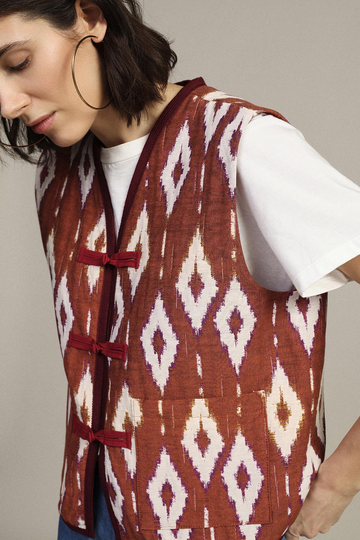 Model wearing rust-red ikat patterned sleeveless vest with toggle closures over white t-shirt, large gold hoop earring