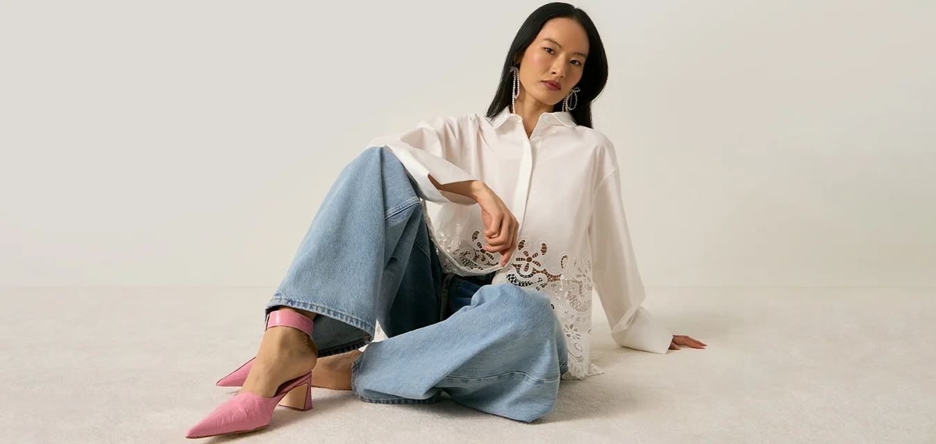 Donna seduta sul pavimento che indossa una camicetta bianca con bordo in pizzo, jeans a gamba larga e scarpe con tacco rosa, su uno sfondo chiaro e semplice.