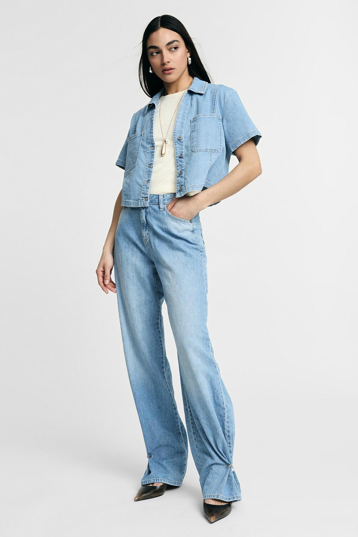 Woman modelling light-blue denim cropped short-sleeve jacket and high-waist wide-leg jeans with cream top and necklace on grey background