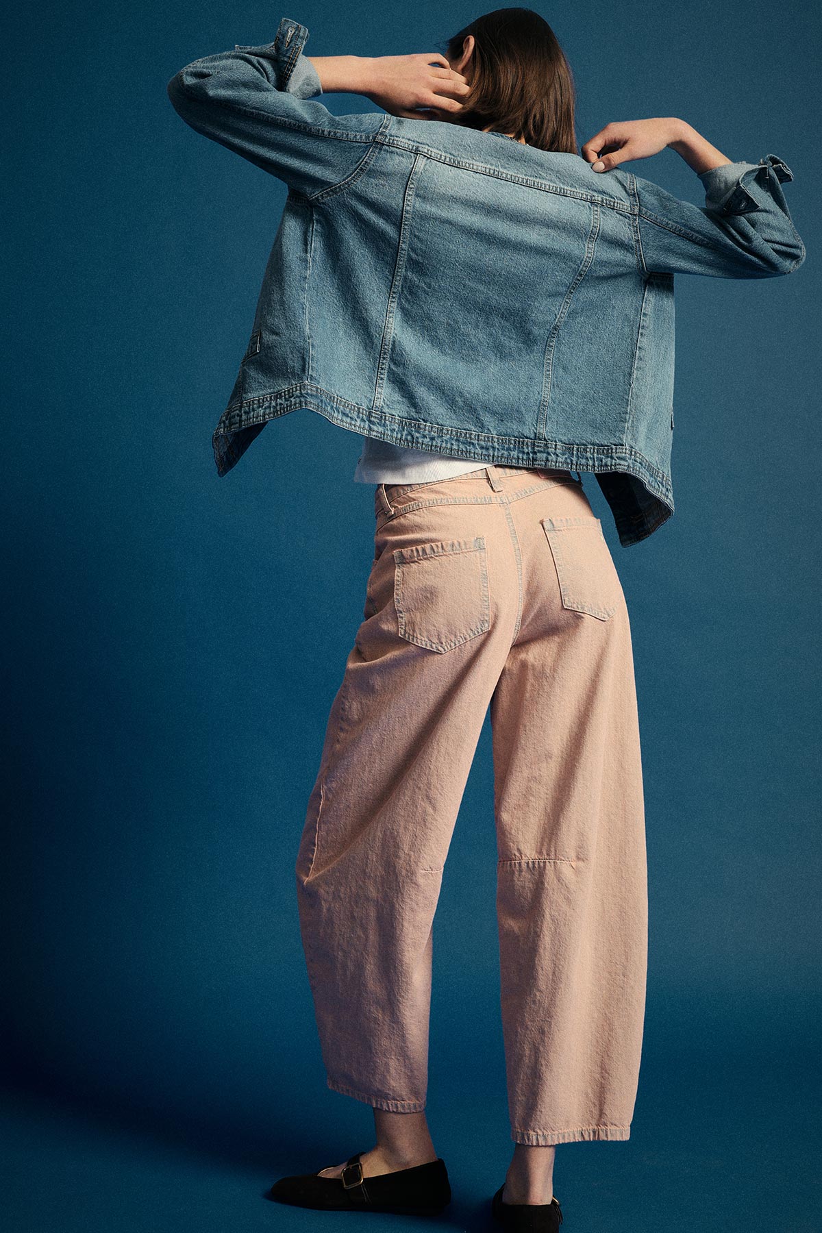 Woman adjusting oversized denim jacket while wearing wide-leg pale pink jeans, back facing camera on blue studio background