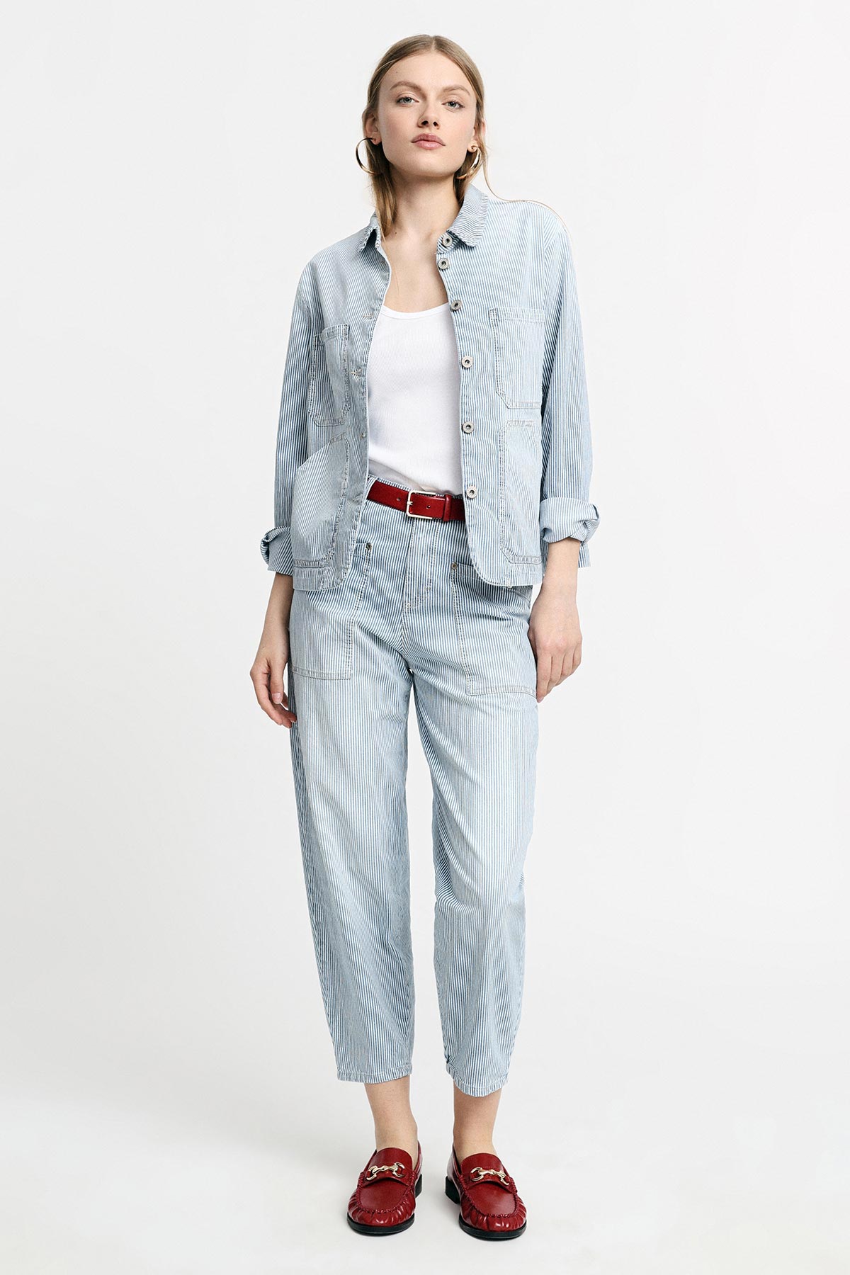 Woman in light blue striped jacket and tapered trousers, white vest, red belt and red loafers in full-length studio pose