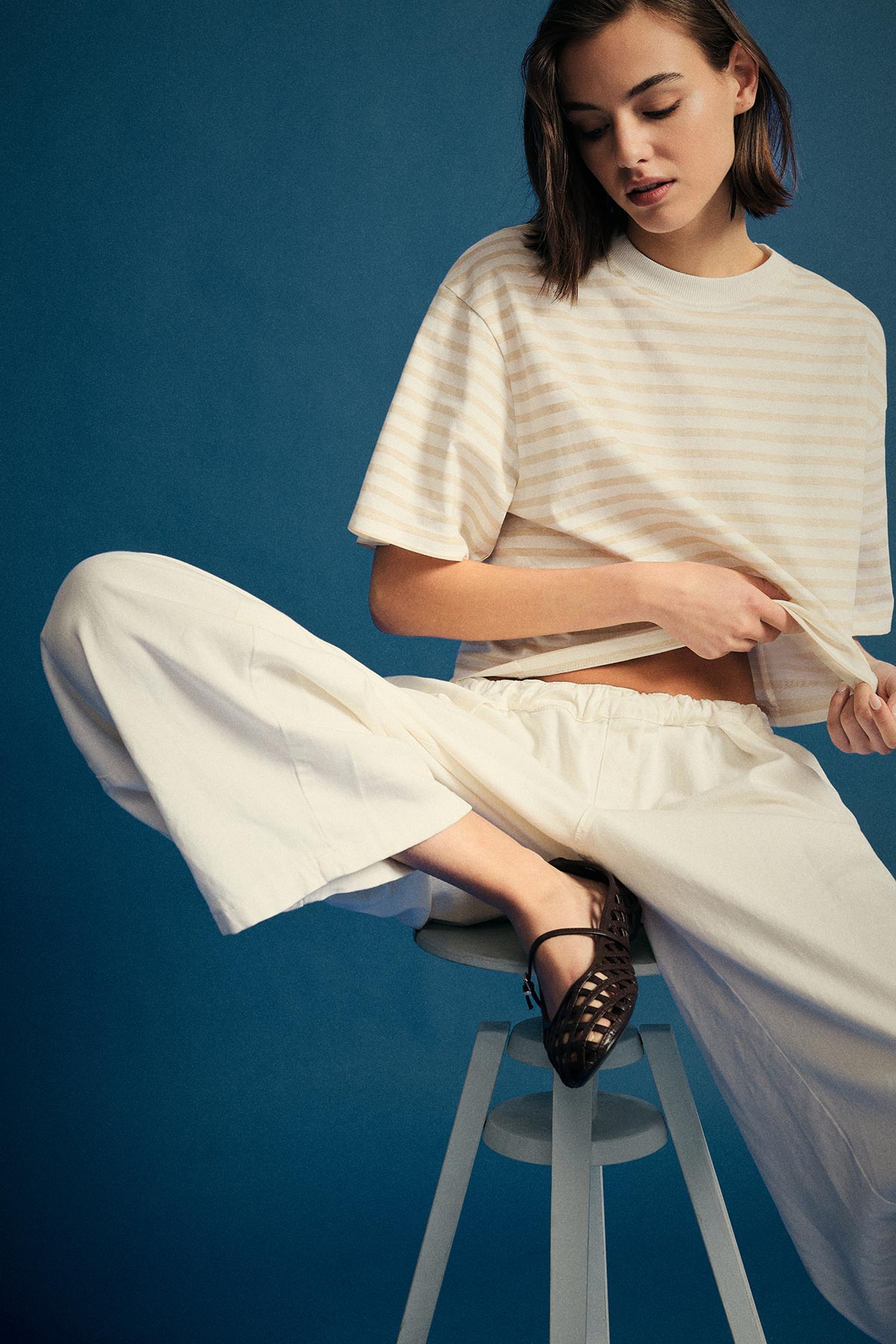 Young woman sitting on a stool against a blue backdrop wearing a cream striped cropped T-shirt, wide white trousers and black woven flats