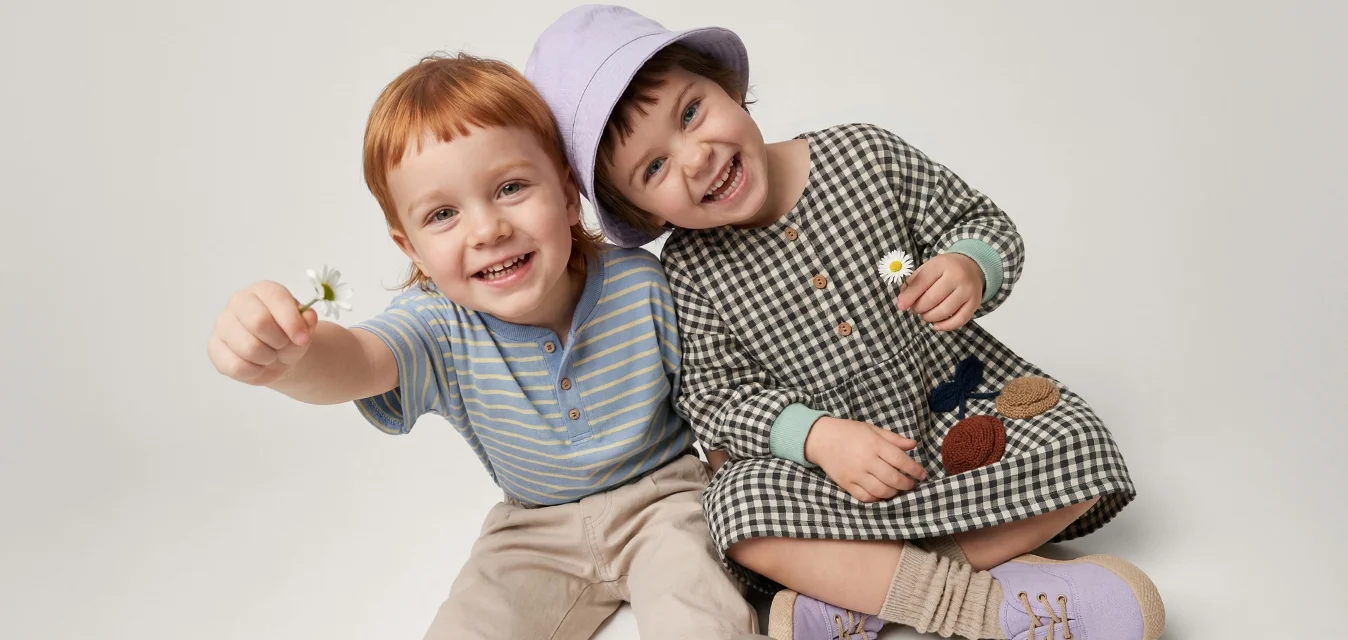 Due bambini sorridenti seduti sul pavimento, tengono piccoli fiori, uno indossa un cappello viola e un vestito a quadretti, l'altro una camicia a righe e pantaloni.