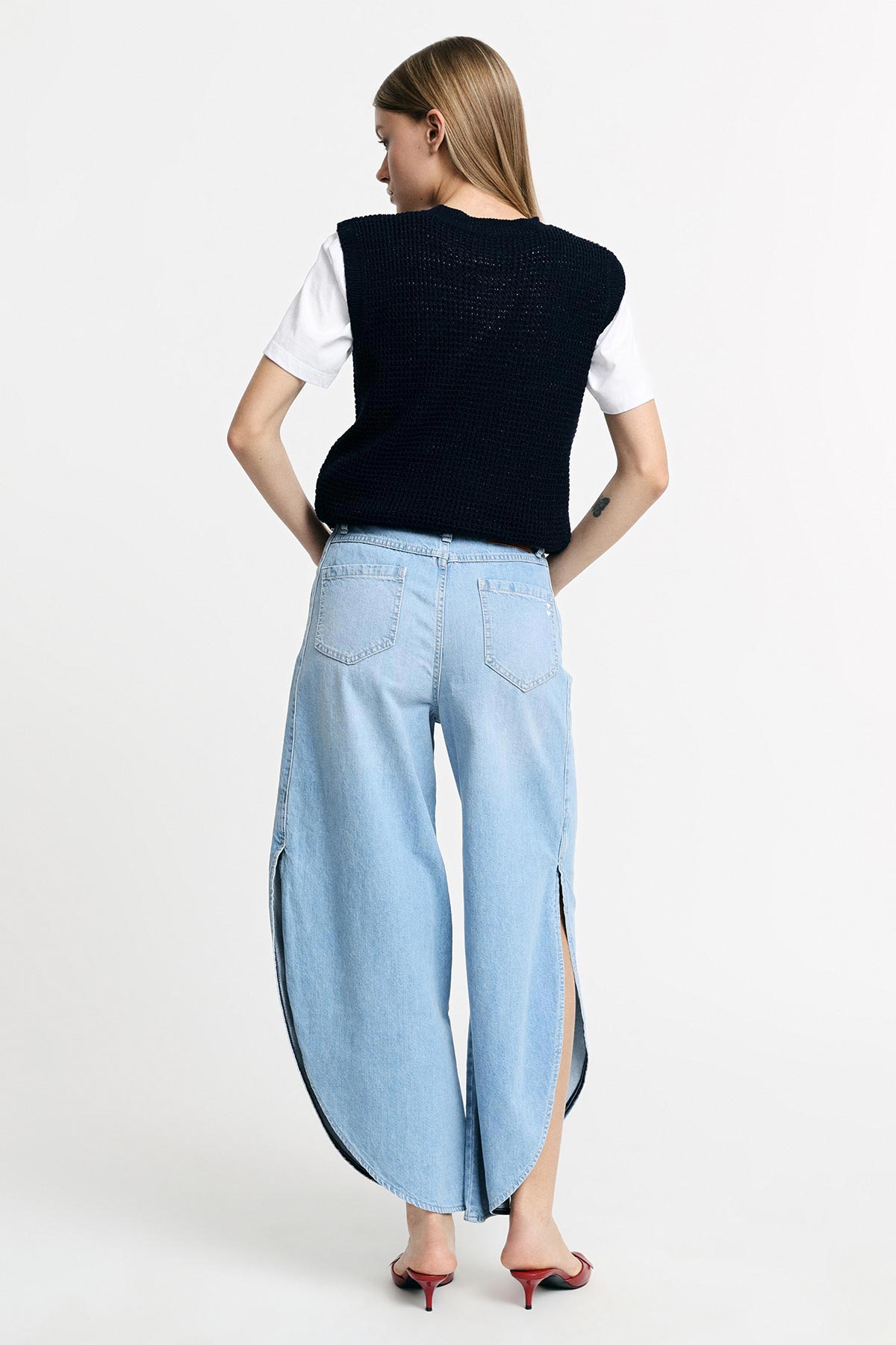 Woman from back wearing light-blue wide-leg jeans with curved side slits, black knit vest over white tee and red heeled sandals