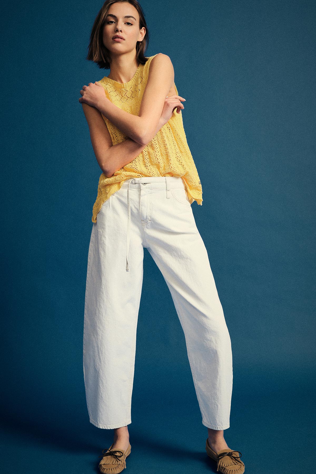 Woman modelling yellow knitted sleeveless top with white wide-leg trousers and tan loafers against a blue studio backdrop