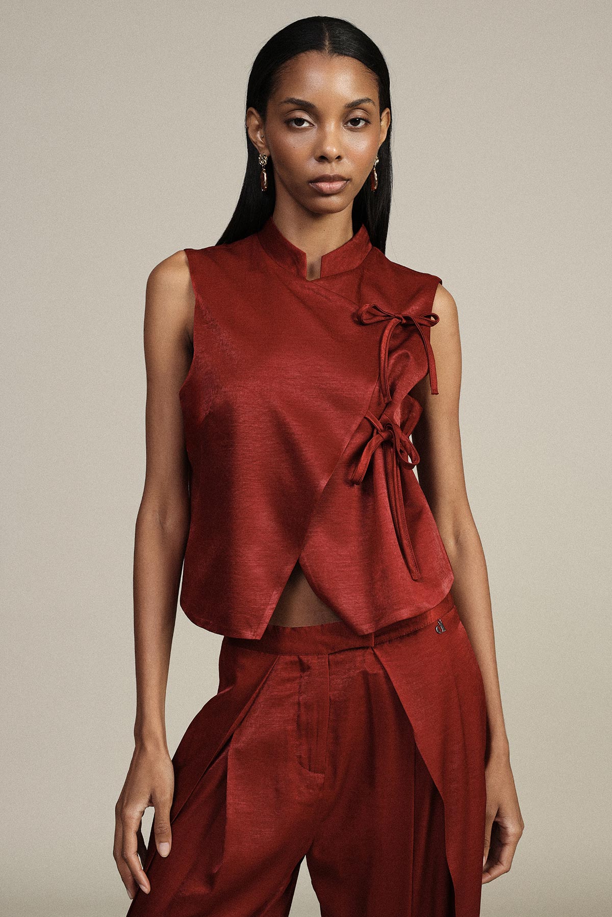 Woman wearing sleeveless red satin top with side bow ties and matching tailored trousers on neutral background