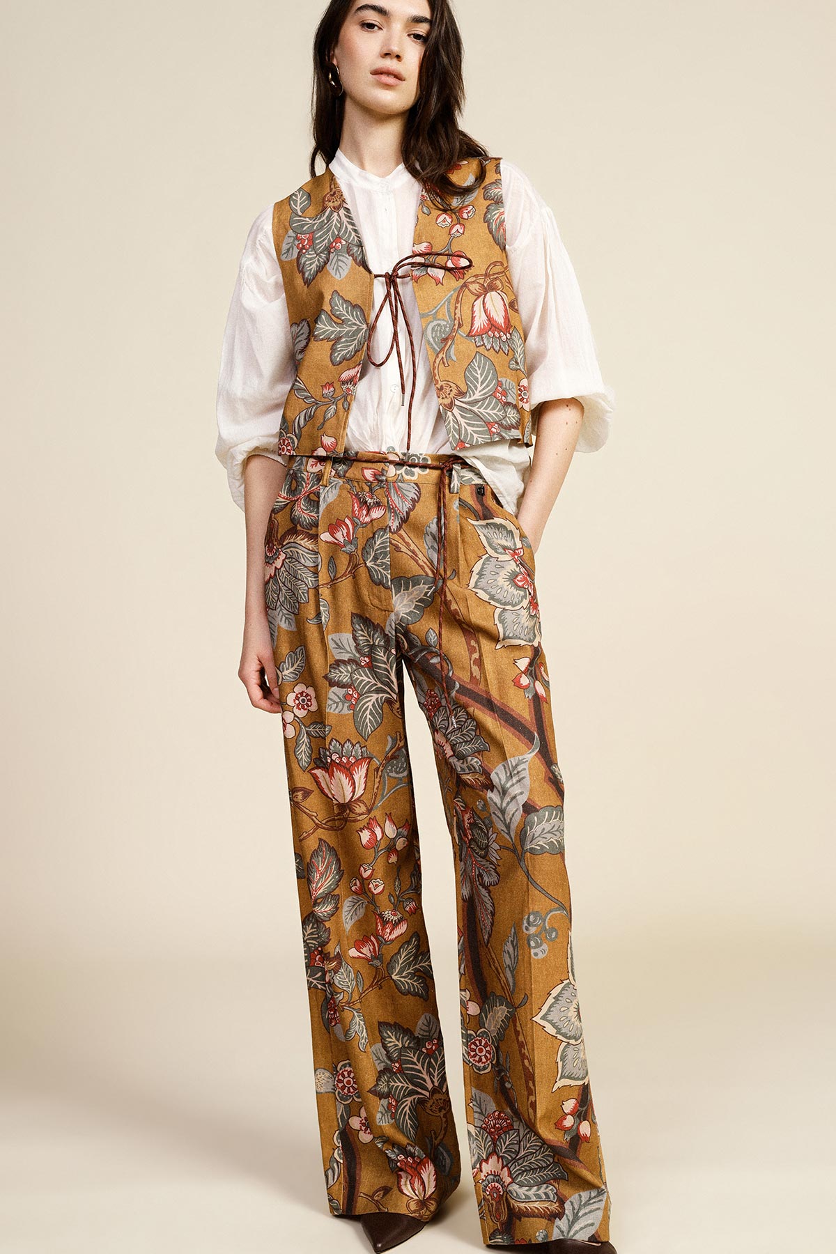 Woman wearing mustard floral vest and matching wide-leg trousers over a white blouse, standing against a neutral background