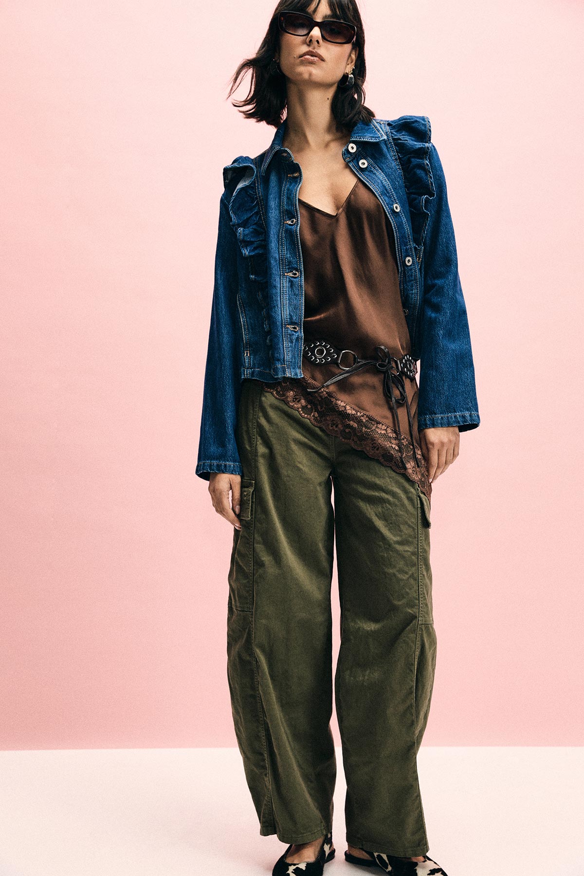 Woman wearing denim ruffle jacket, brown silk camisole with lace, olive wide trousers and belt, sunglasses, pink background