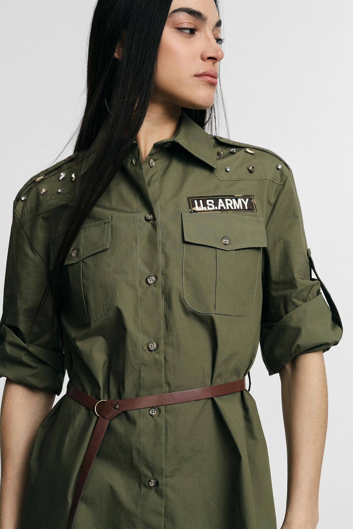 Young woman in olive green U.S. Army-style shirt with studded shoulders, rolled sleeves and brown belt looking sideways