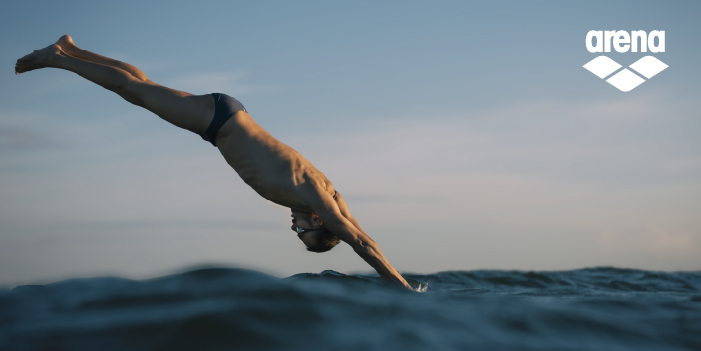 A swimmer diving into open water at sunset wearing Arena swim briefs, ocean surface and horizon