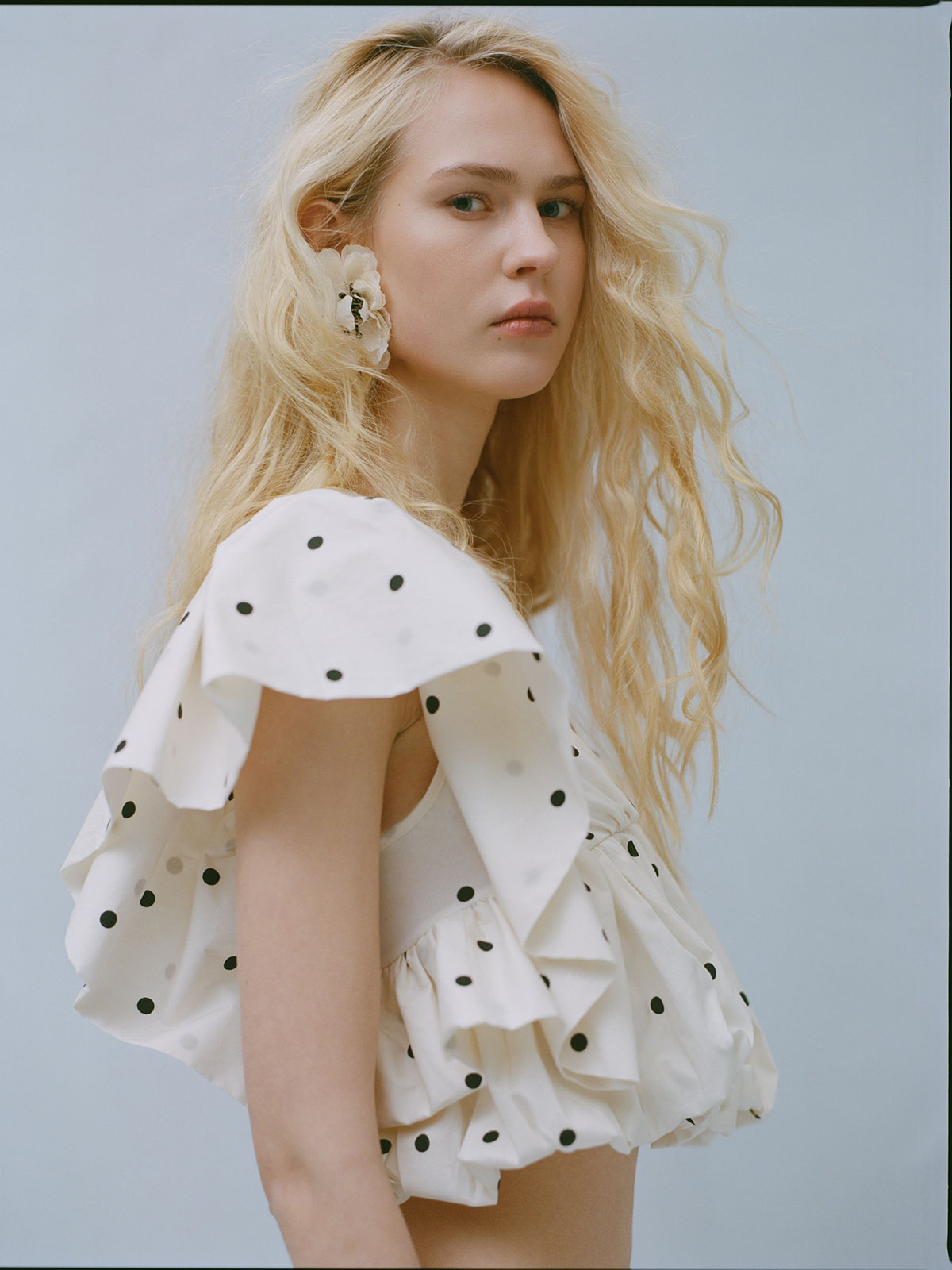 Side-profile of a young blonde woman wearing a white polka-dot ruffled top and large flower earring against a pale background