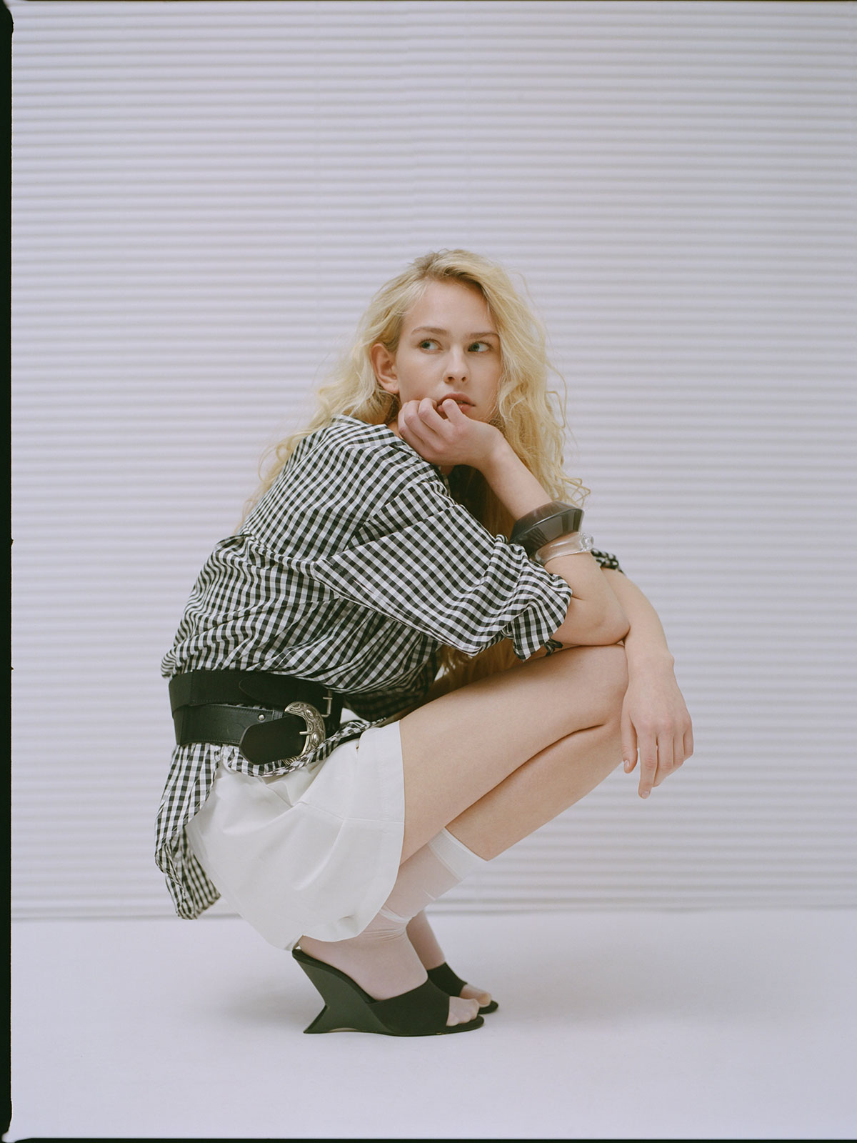Blonde woman in black-and-white gingham shirt, white skirt and black wedge heels, squatting and gazing sideways against pale backdrop