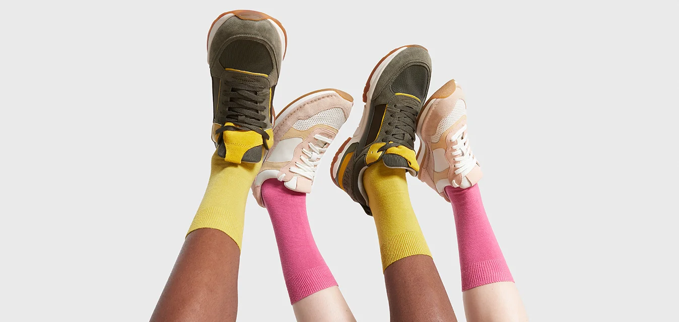 Due paia di sneaker: una verde oliva con accenti gialli, l'altra beige con rete bianca. Calzini gialli e rosa brillanti visibili.