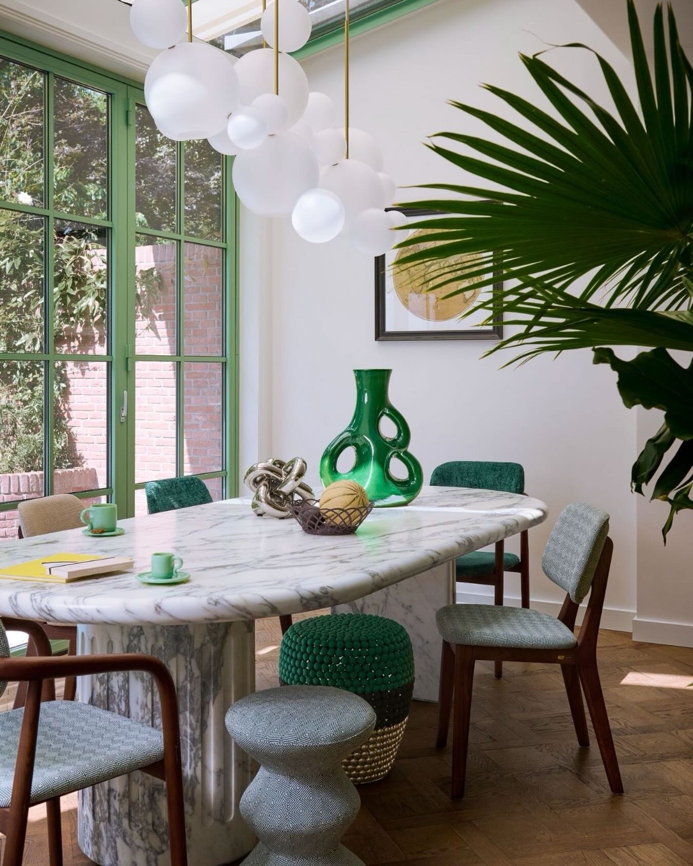 Bright dining room with oval marble table, green glass vase and accents, globe pendant lights and large green-framed window