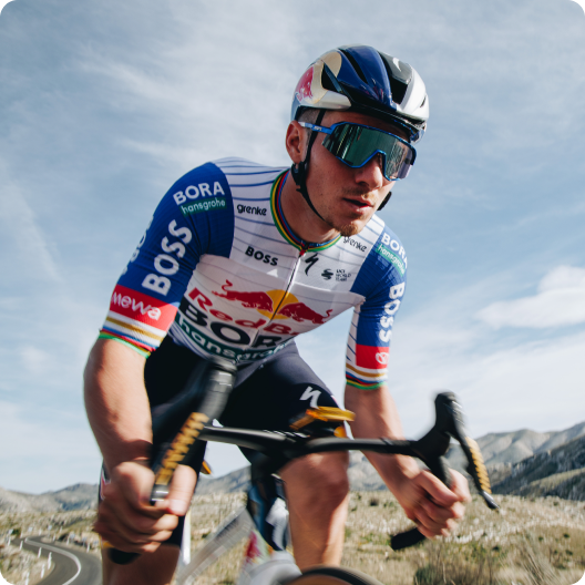 Cyclist in professional Bora‑Red Bull team kit wearing helmet and sunglasses riding a road bike on a mountain road