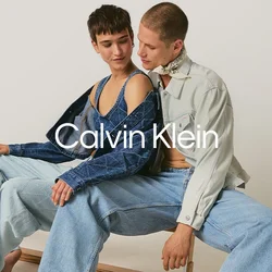 Due giovani adulti scalzi con indosso abbigliamento in denim sono seduti vicini su una panchina di legno davanti a uno sfondo semplice.