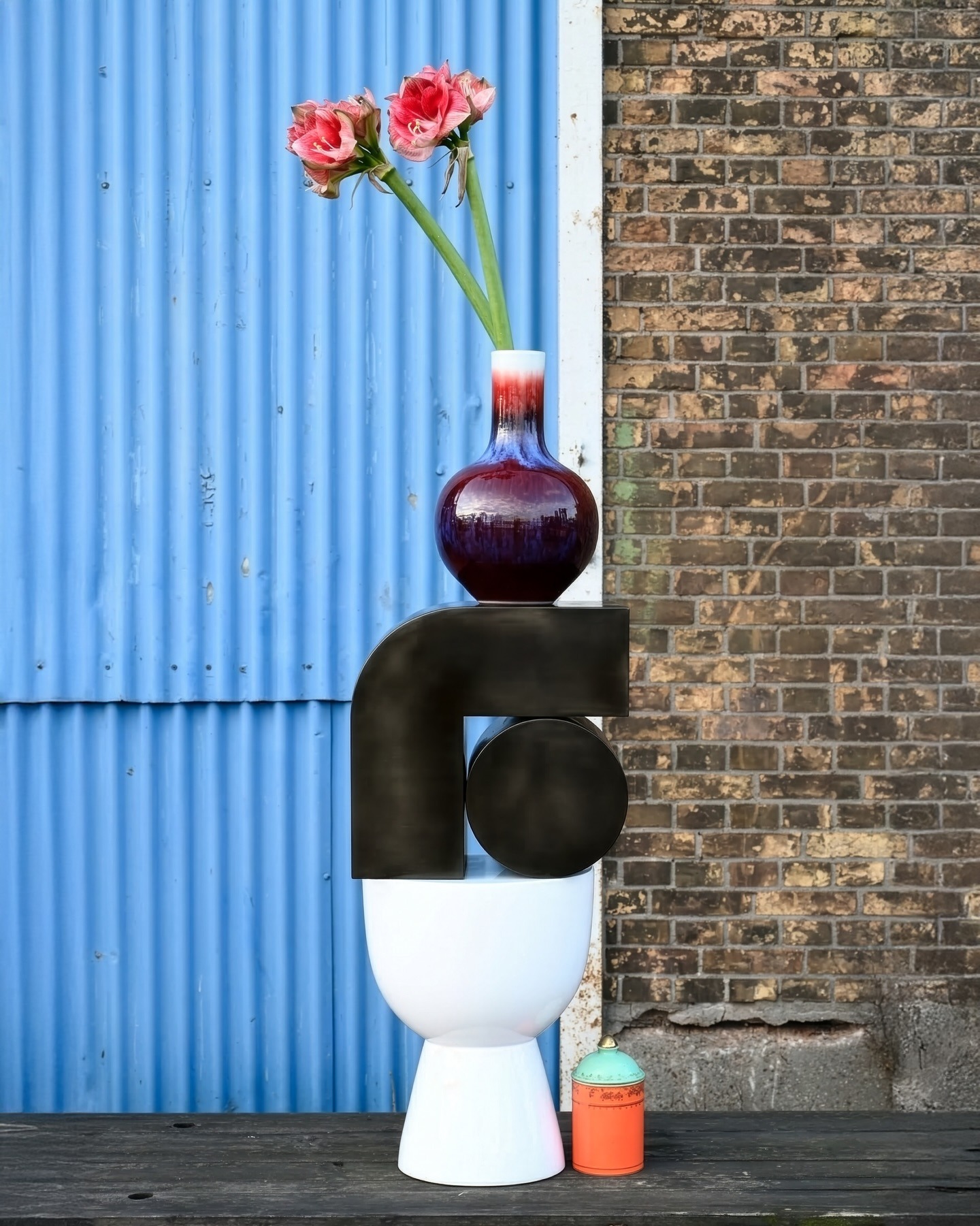 Modern still-life: red glass vase with pink amaryllis on stacked sculptures against blue corrugated metal and brick wall