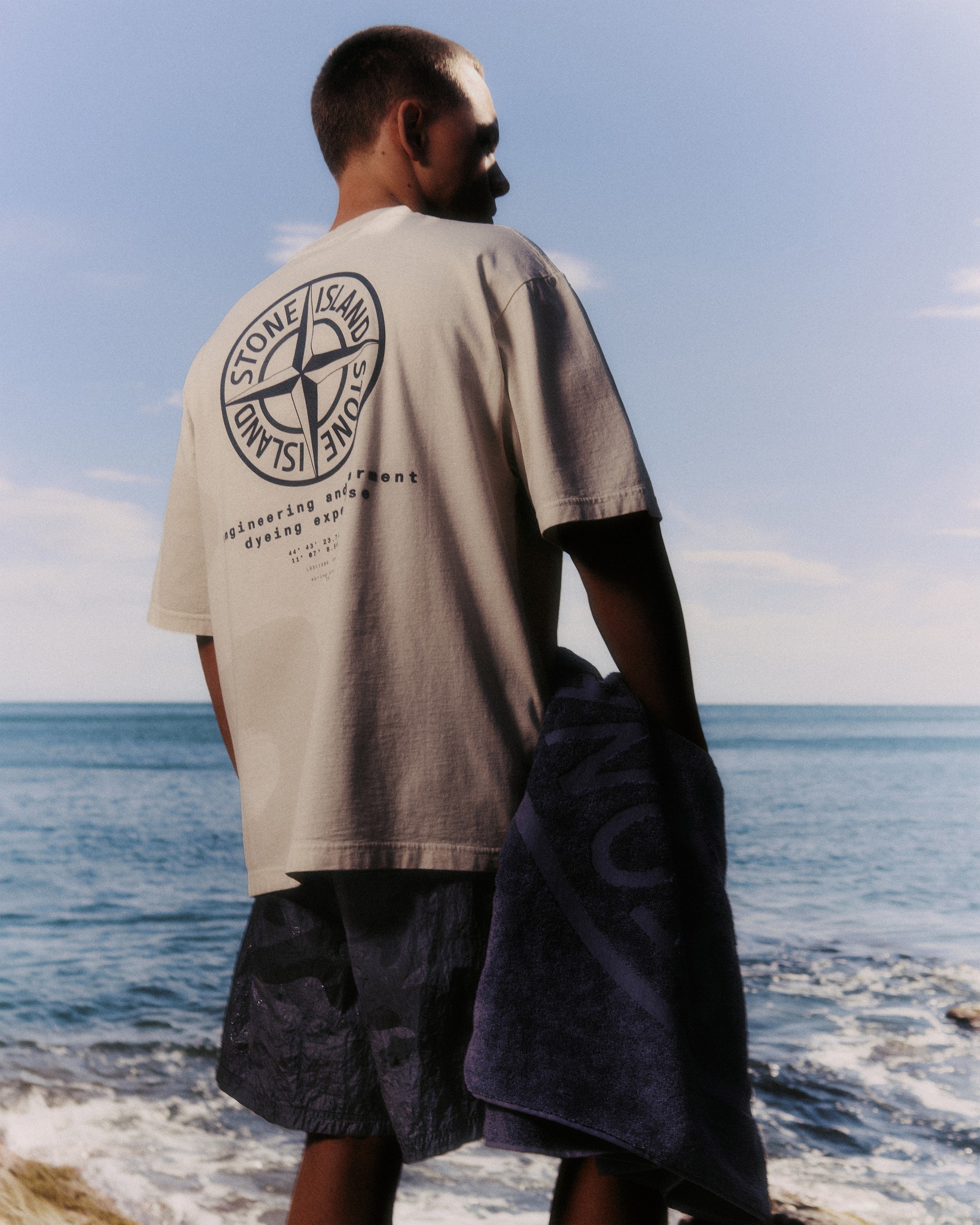 Man on rocky beach facing the sea, wearing beige Stone Island T-shirt with back logo and holding navy towel
