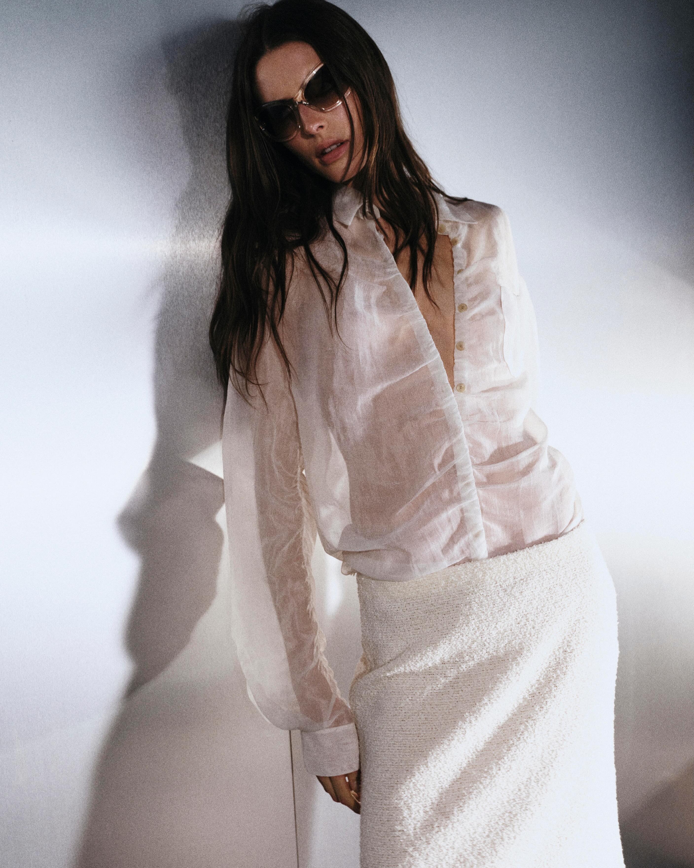 Fashion model wearing a sheer white blouse, textured cream pencil skirt and sunglasses, leaning against a wall in soft studio light