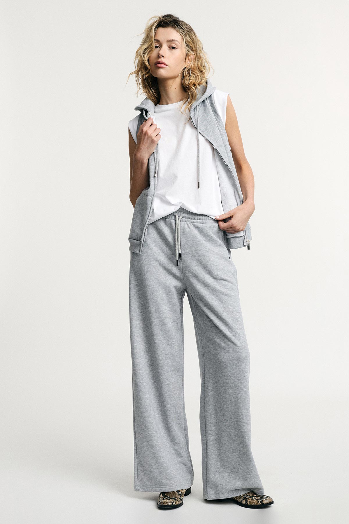 Woman in grey wide-leg sweatpants, sleeveless grey zip hoodie and white sleeveless top, patterned shoes, neutral studio background