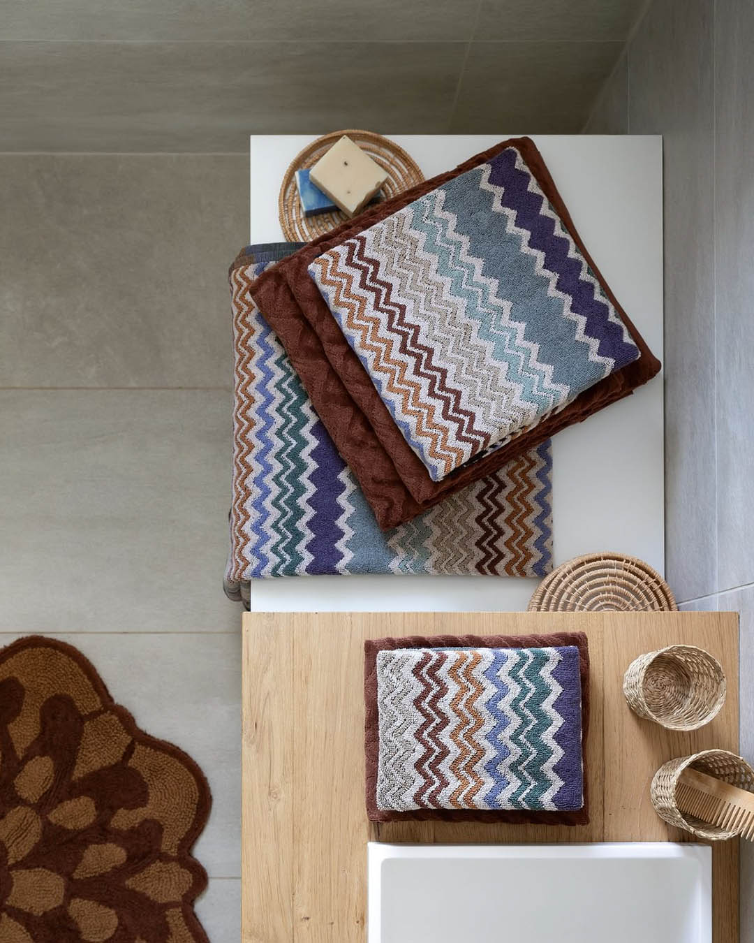 Stack of folded zigzag-patterned bath towels in blue, purple and brown on a wooden bathroom counter with wicker baskets and soap