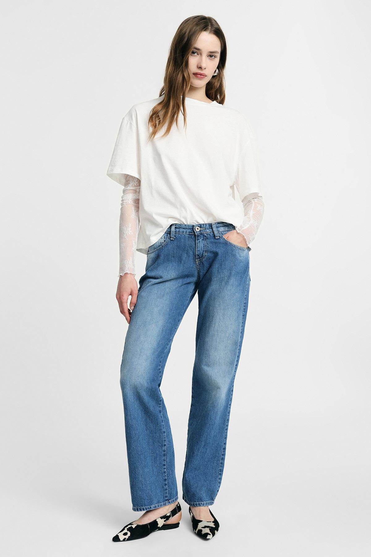 Young woman modelling white oversized T-shirt with lace long sleeves and straight-leg blue jeans, black-and-white flats on plain background