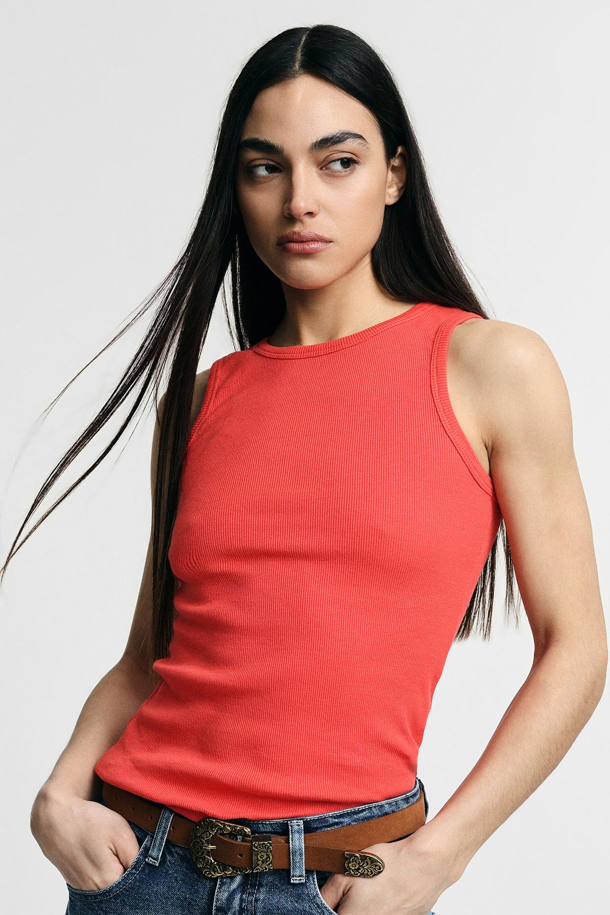 Woman in red ribbed sleeveless top and jeans with decorative brown belt, long dark hair, hands in pockets