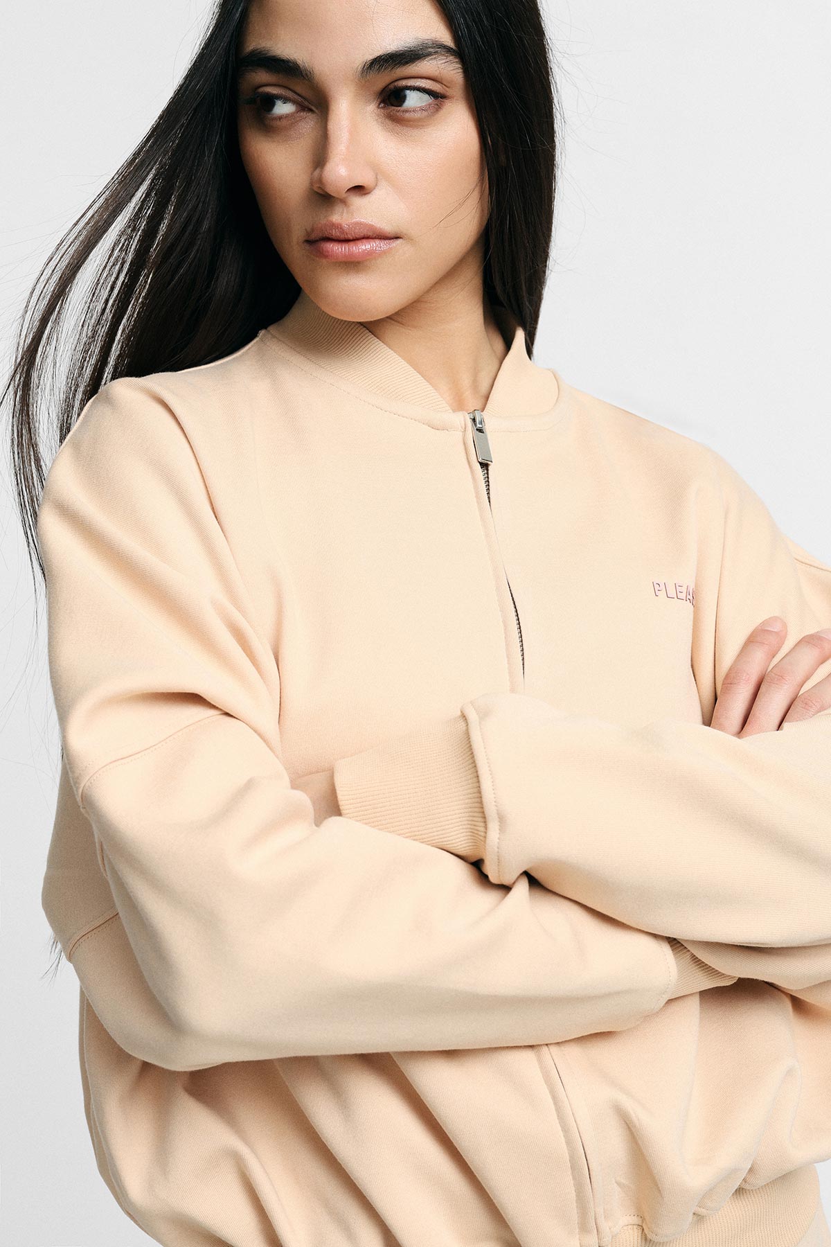 Young woman model with long dark hair wearing a beige zip-up bomber jacket, arms crossed, looking sideways