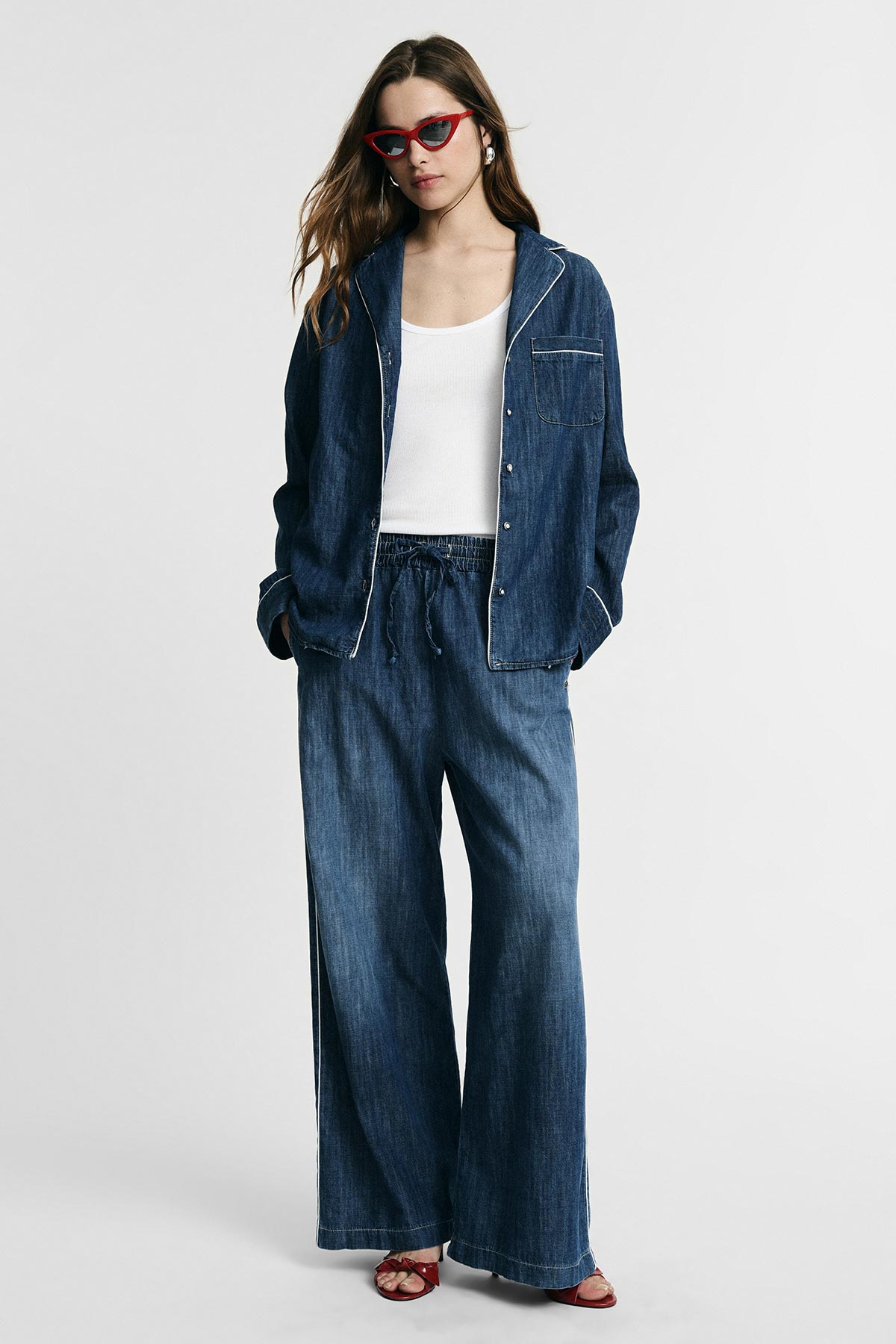Woman wearing denim pyjama-style jacket and wide-leg jeans with white vest top, red cat-eye sunglasses and red sandals