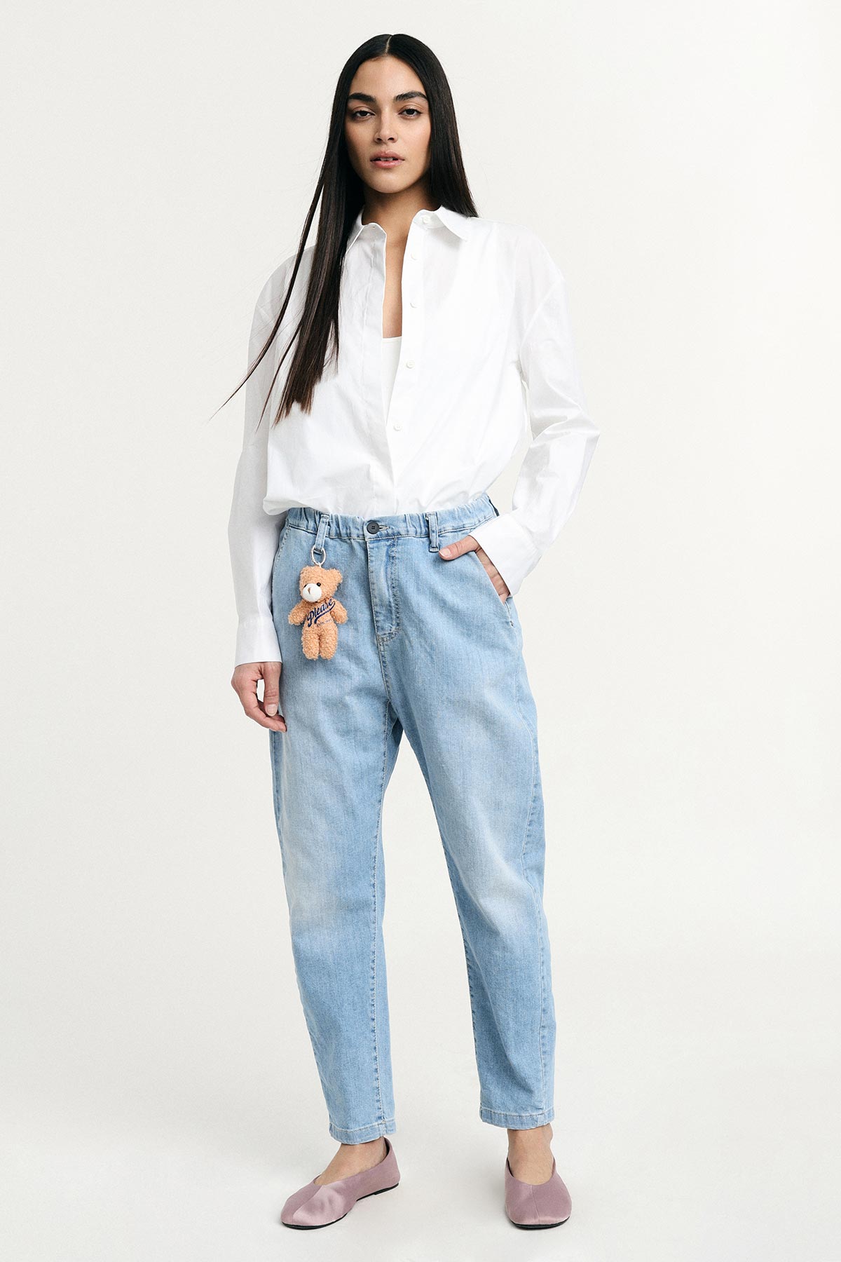 Young woman standing in white shirt and light-blue high-waist jeans with teddy keyring, mauve flats, hands in pockets