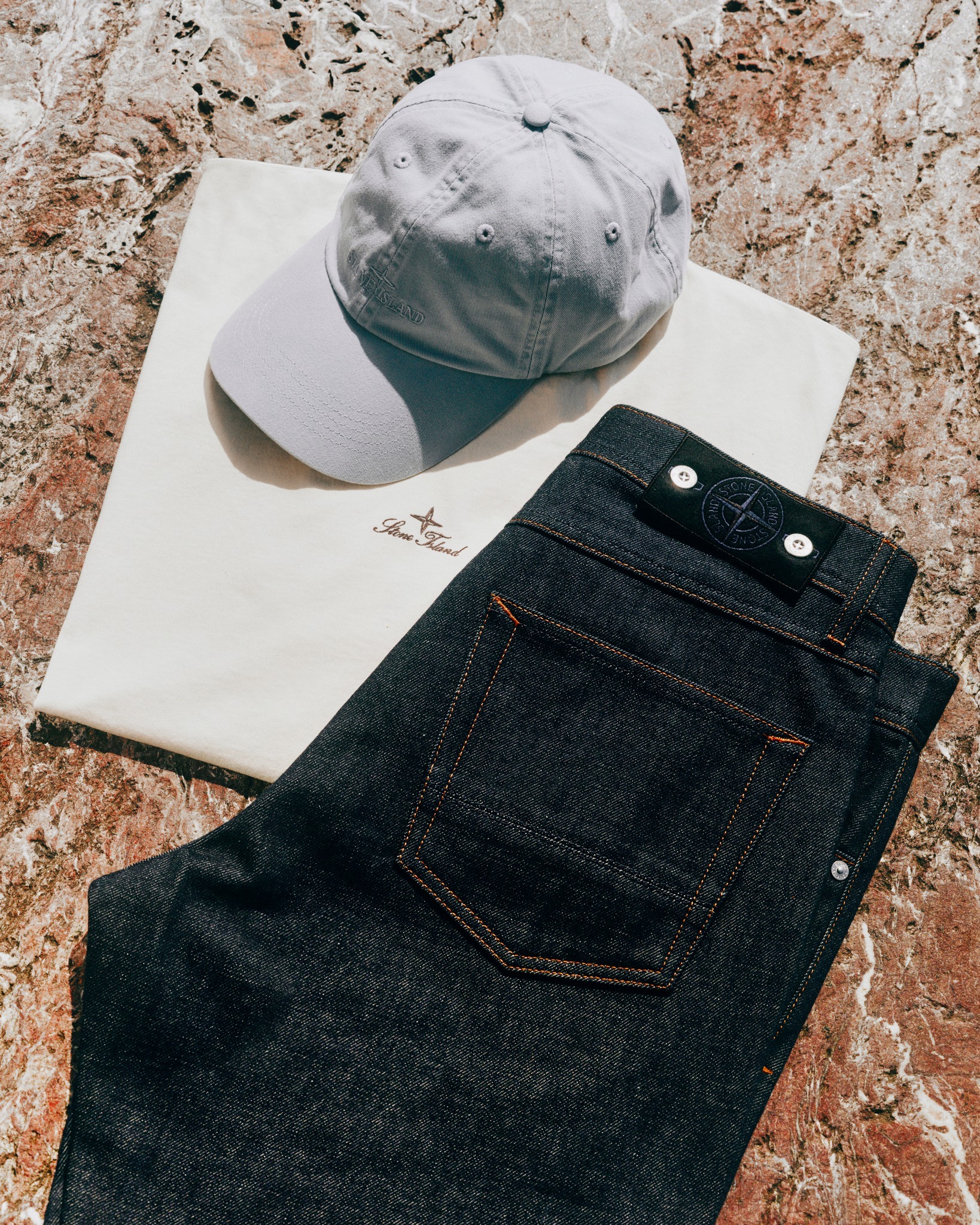 Flatlay of dark denim jeans with leather brand patch, cream t-shirt with small logo and light grey baseball cap on marble surface