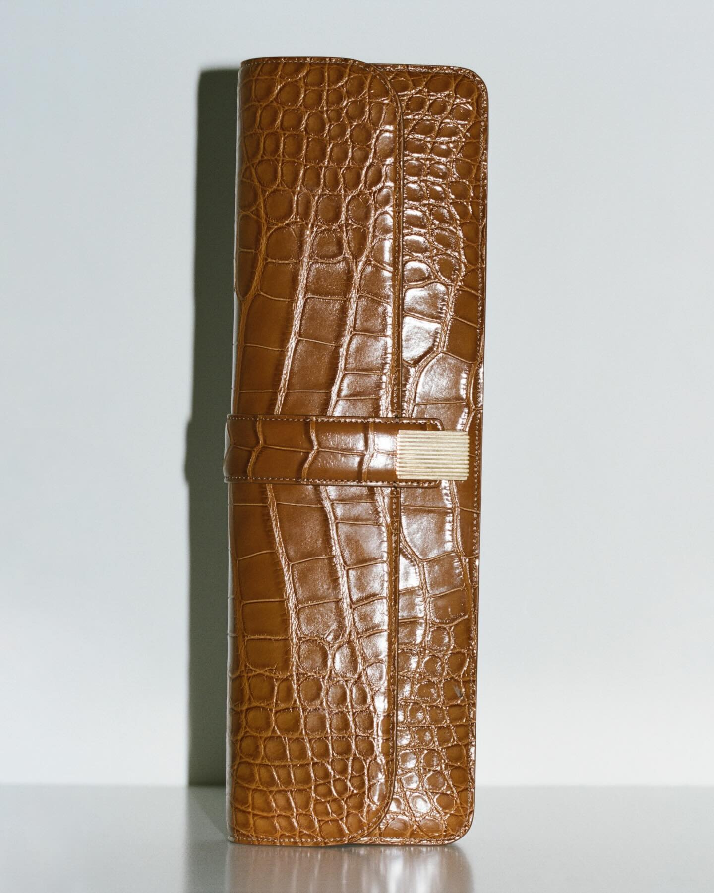 Brown crocodile‑embossed leather roll-up jewellery case with gold clasp, vertical on pale background for luxury travel storage