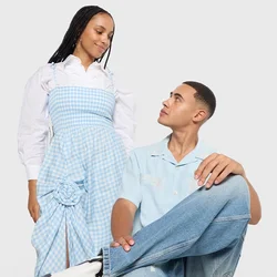 Donna con un vestito a quadri blu in piedi accanto a un uomo seduto su una sedia rossa, che indossa una camicia azzurro chiaro e jeans, entrambi si guardano.
