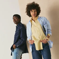 Due persone stanno appoggiate in modo informale a un muro; una indossa una camicia di jeans blu e jeans, l'altra una camicia azzurro chiaro, una maglietta gialla e jeans.