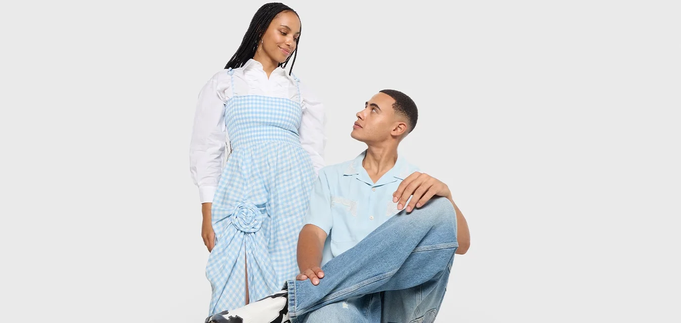 Donna con un vestito a quadri blu in piedi accanto a un uomo seduto su una sedia rossa, che indossa una camicia azzurro chiaro e jeans, entrambi si guardano.