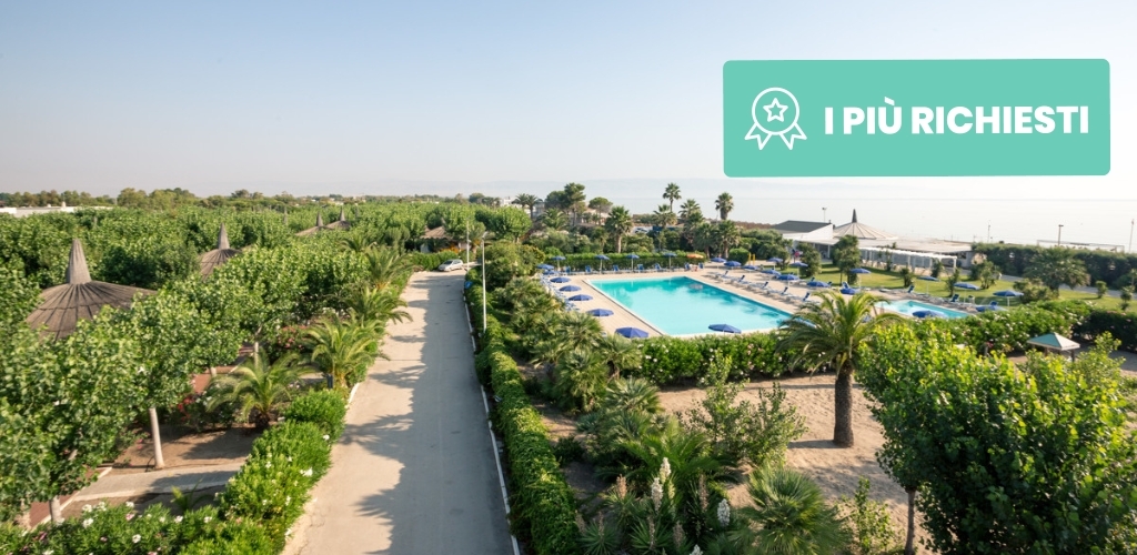 Aerial view of coastal beach resort with outdoor pool, palm trees, pathway and sea in background, 'I PIÙ RICHIESTI' badge