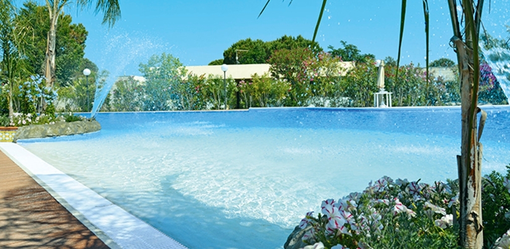 Resort swimming pool with clear blue water and decorative fountains, palm trees and flowering shrubs under a bright blue sky