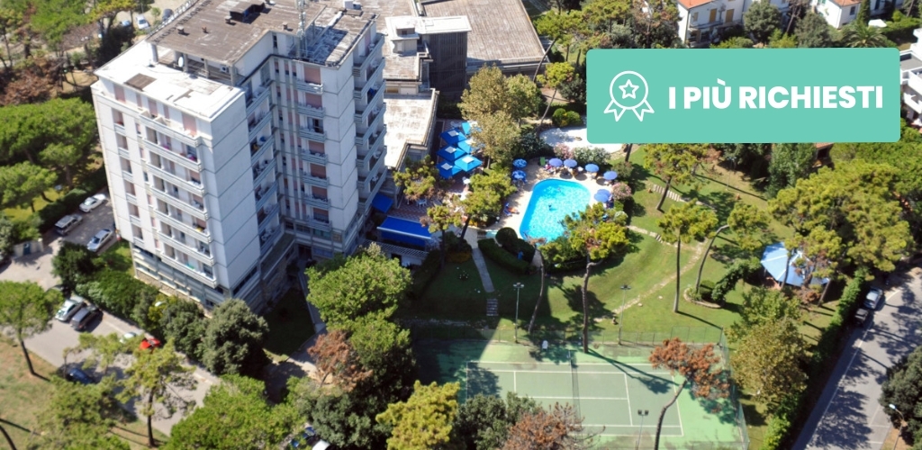 Aerial view of a multi-storey hotel surrounded by trees, outdoor swimming pool with umbrellas and a tennis court in landscaped gardens