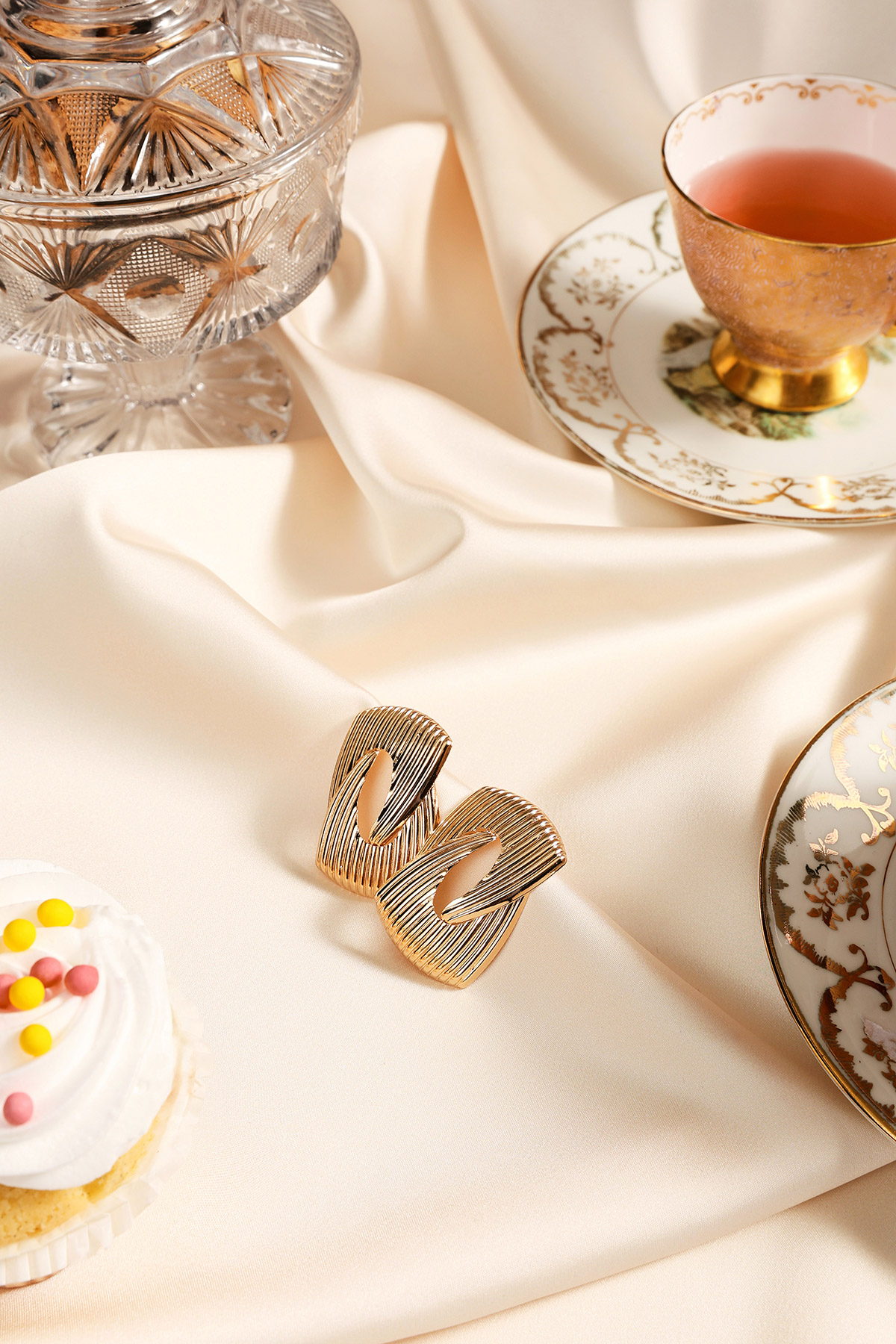 Pair of textured gold clip-on earrings on cream satin beside vintage china teacup, crystal candy jar and decorated cupcake