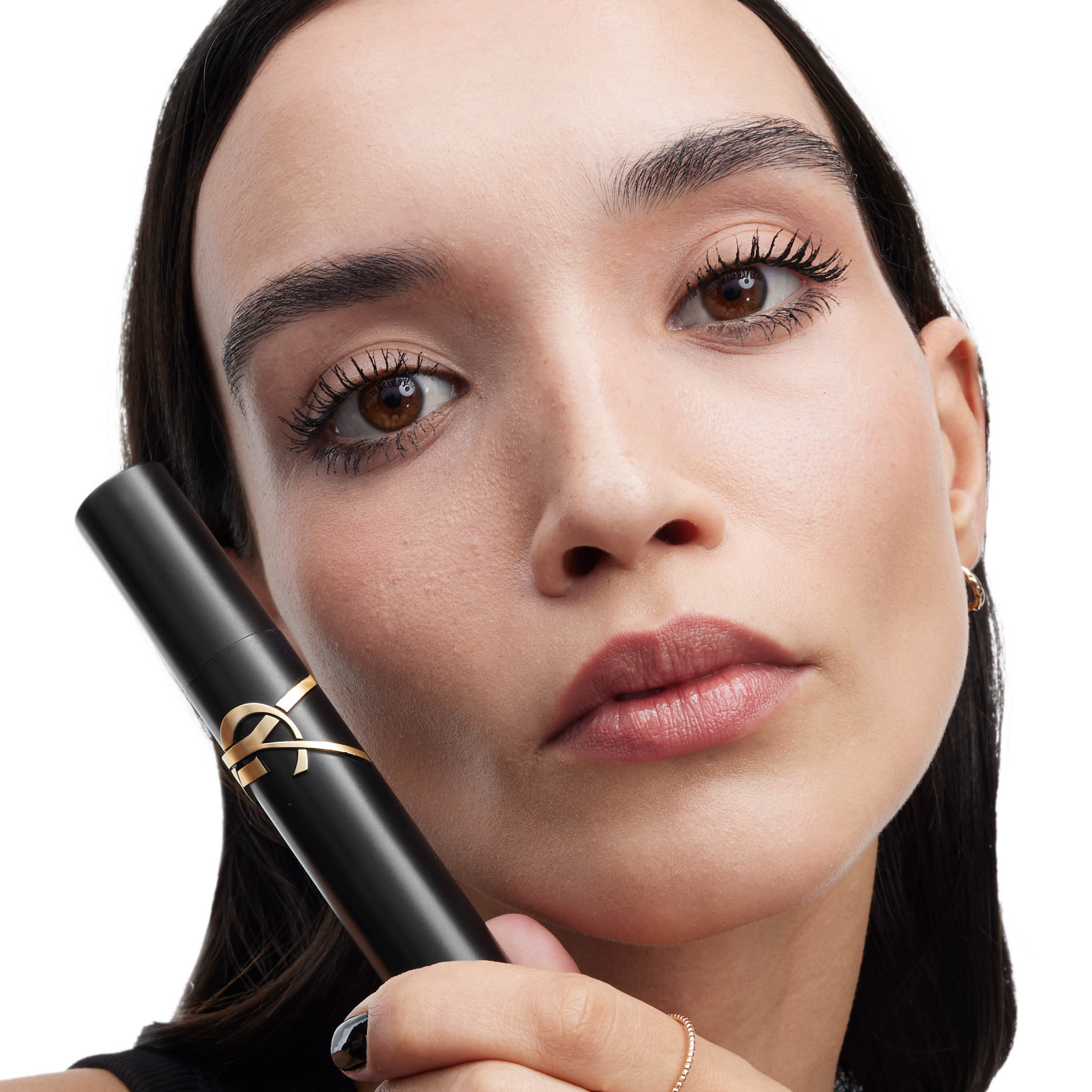 Primo piano di una modella bruna con ciglia intense e definite che tiene il mascara YSL Beauty Lash Clash nel suo iconico packaging nero e dorato su sfondo bianco