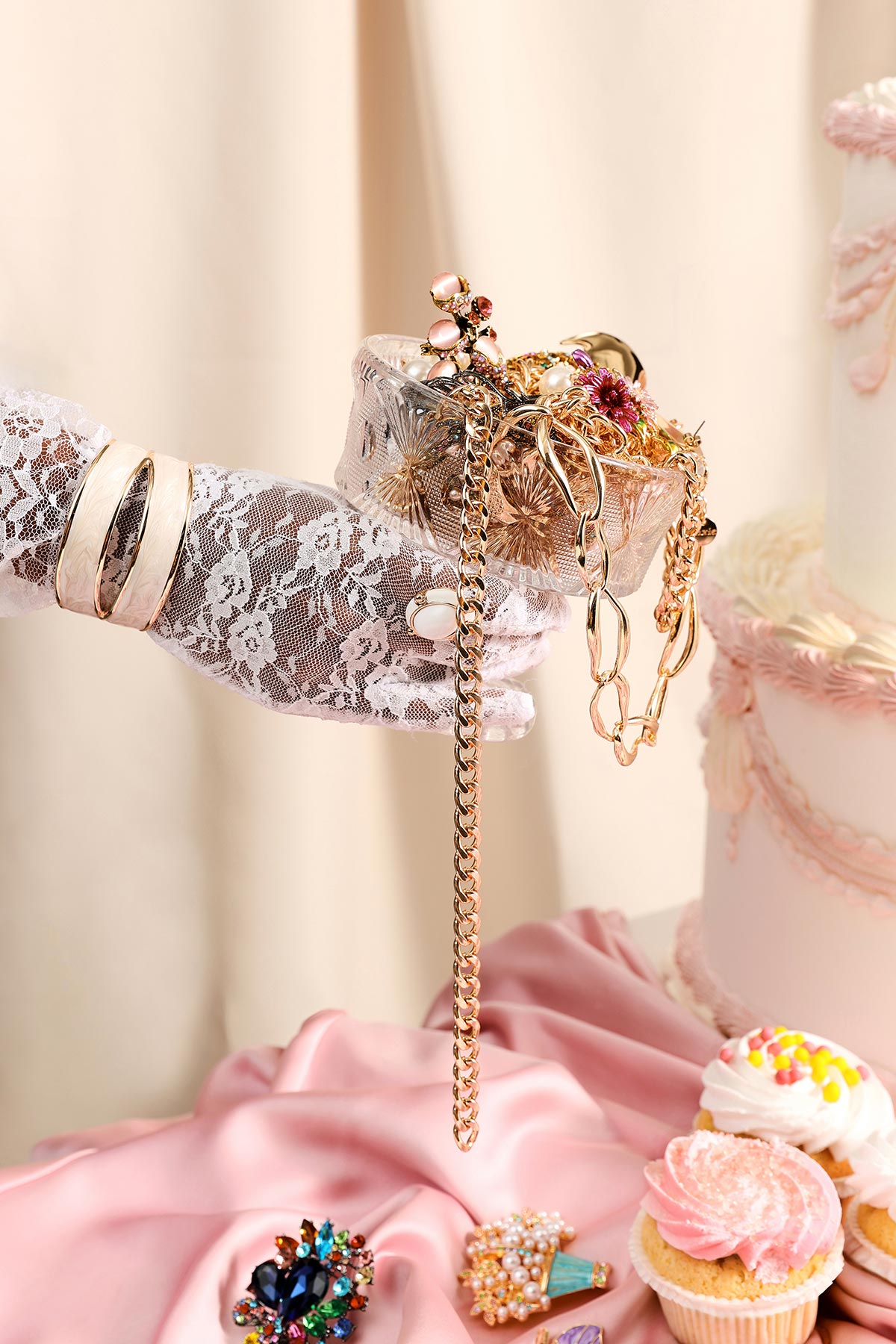 Lace-gloved hand holding crystal dish of gold jewellery and chains beside pink frosted cake and cupcakes on satin