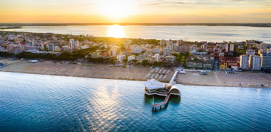 Aerial view of a sunlit seaside town at sunset with rows of beach sunbeds, promenade and a curved pier restaurant over calm sea