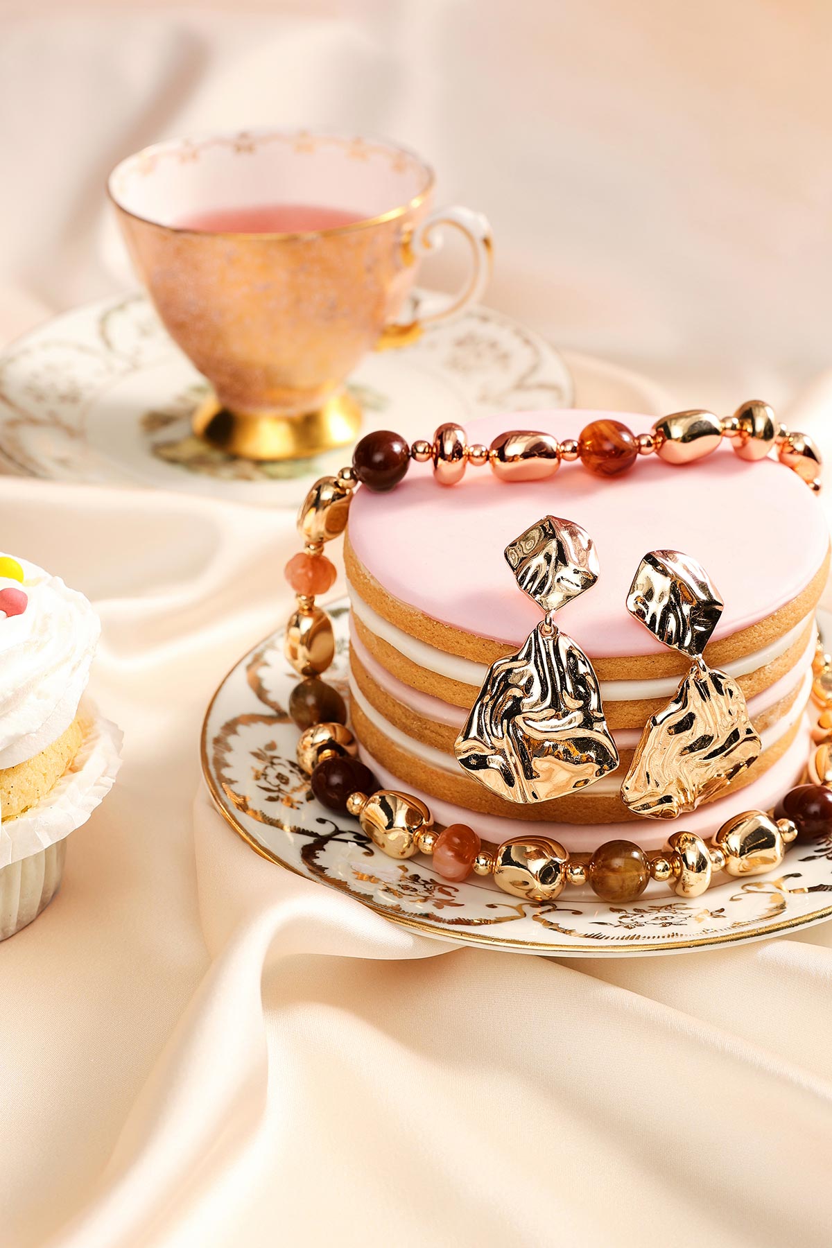 Gold abstract drop earrings and beaded necklace draped over a pink layered macaron cake on vintage china plate with a teacup