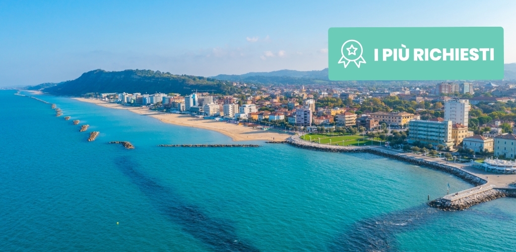 Aerial view of sunlit seaside town with turquoise water, sandy beach, breakwaters and promenade, green 'I PIÙ RICHIESTI' badge