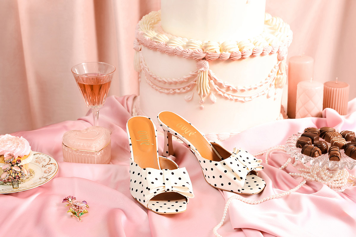 White polka-dot bow heeled mules on pink satin beside a tiered cake, chocolates, pearl necklace and a glass of rosé