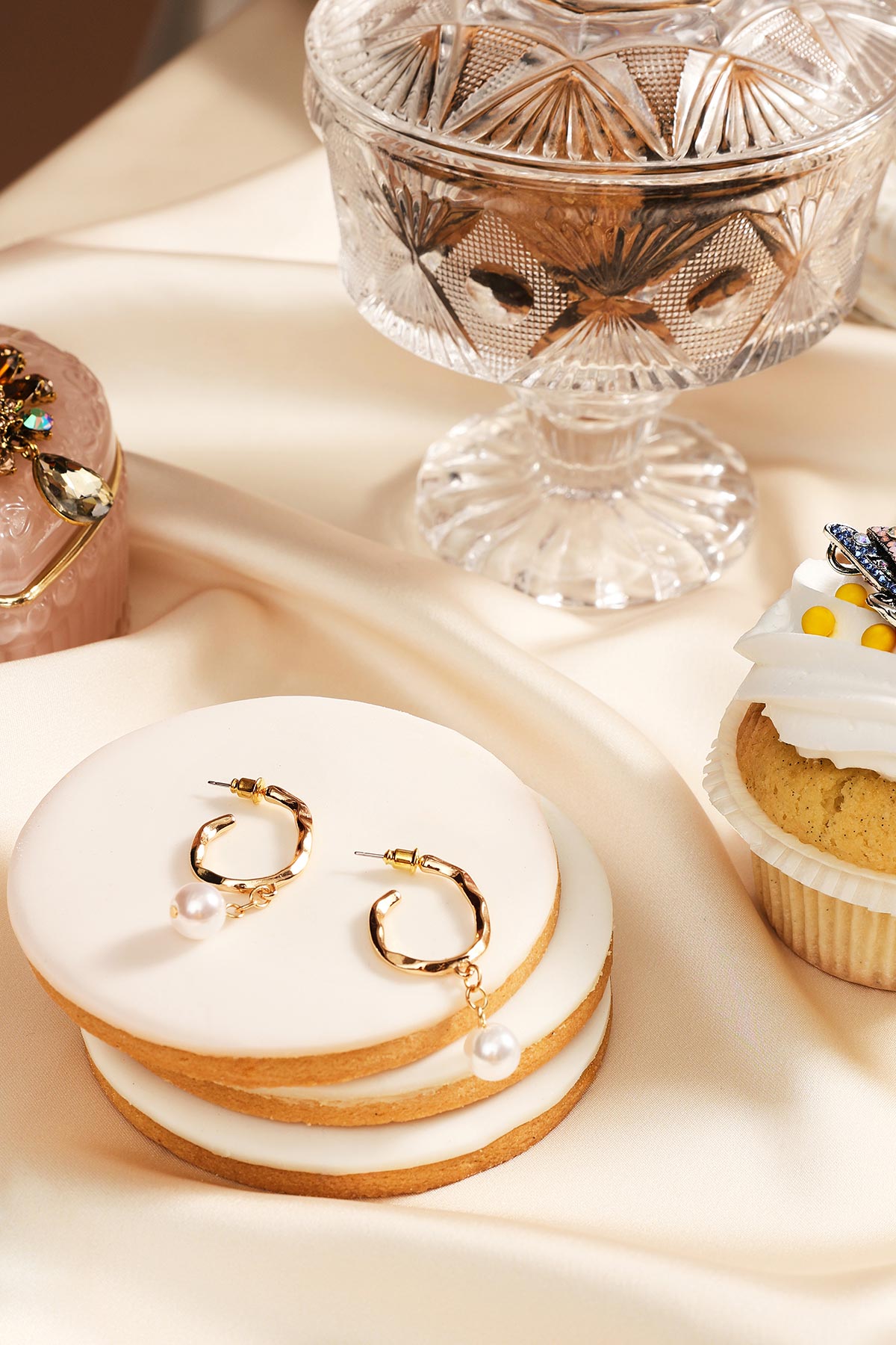 Gold hoop earrings with dangling pearls displayed on stacked white-frosted biscuits, crystal dish and cupcake on satin