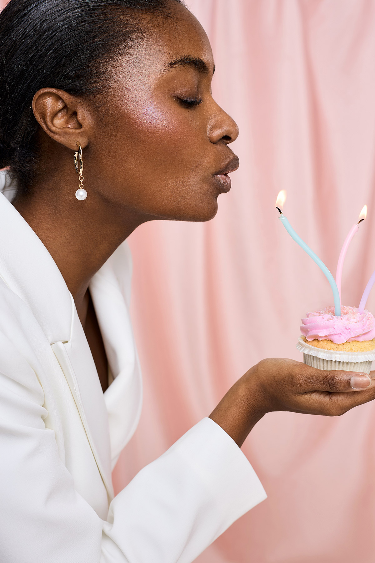 Woman in white blazer blowing out two lit candles on a pink-frosted cupcake against a pink backdrop